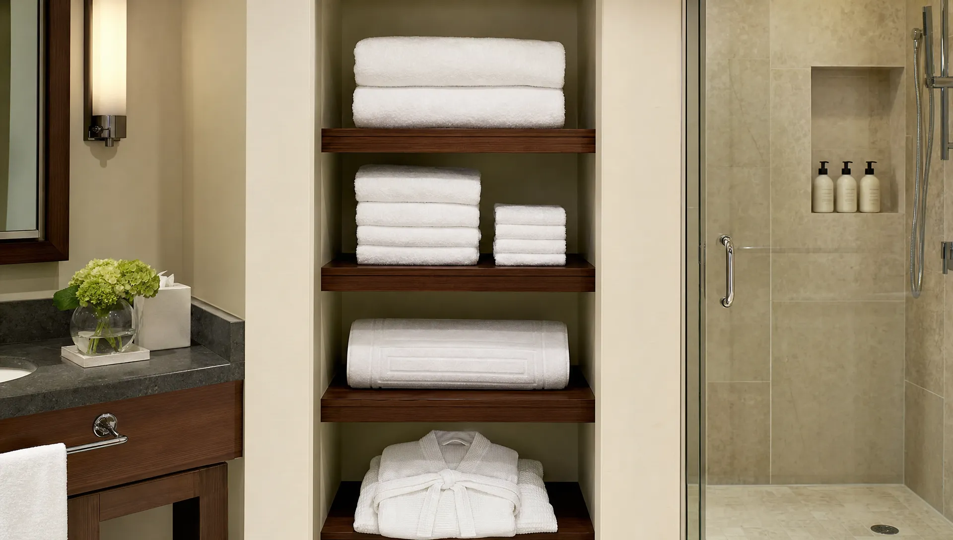 A hotel bathroom shelf holds bath towels, hand towels, washcloths, a bath mat, and a robe beside a vanity and shower area.